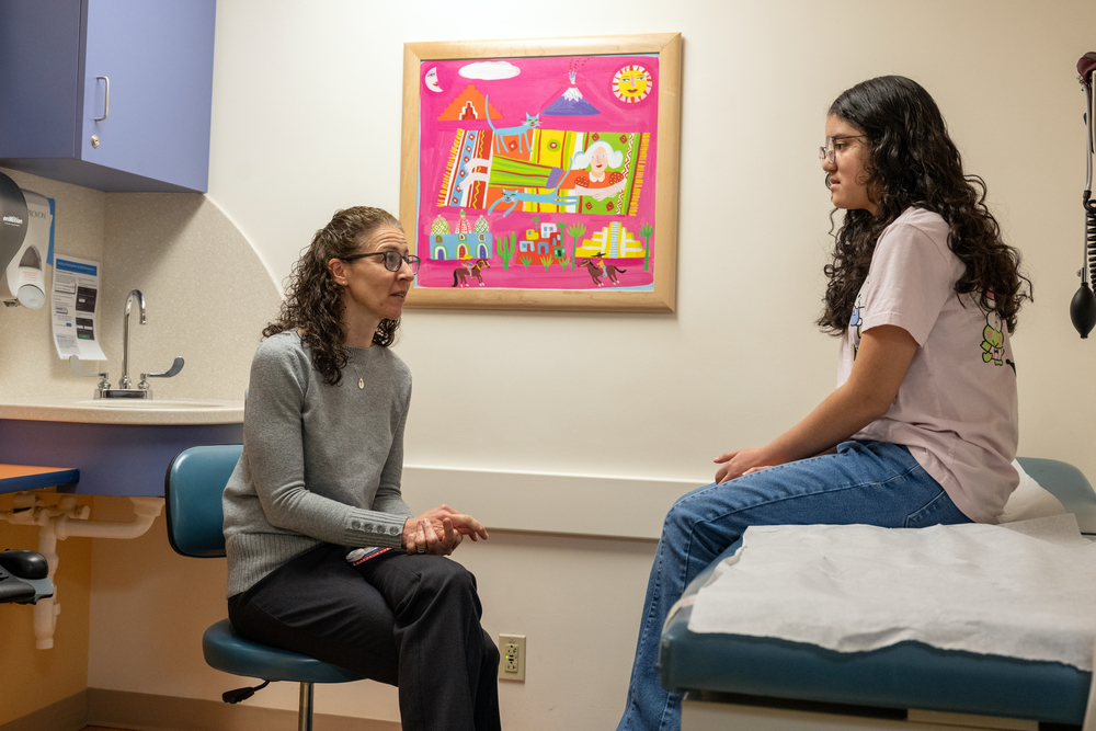 Female clinician sitting on chair looks at teenage female patient sitting on exam bench