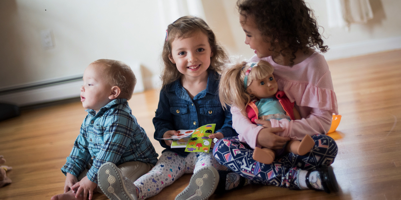 Three children play on hardwood floor. One holds a baby doll. A girl smiles toward camera.