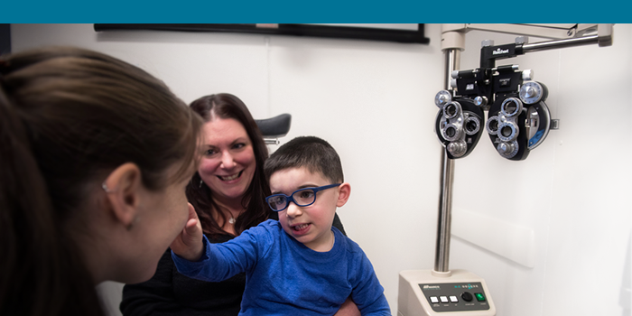 Boy wearing glasses sits on mom's lap. Boy pokes at nose of clinician.