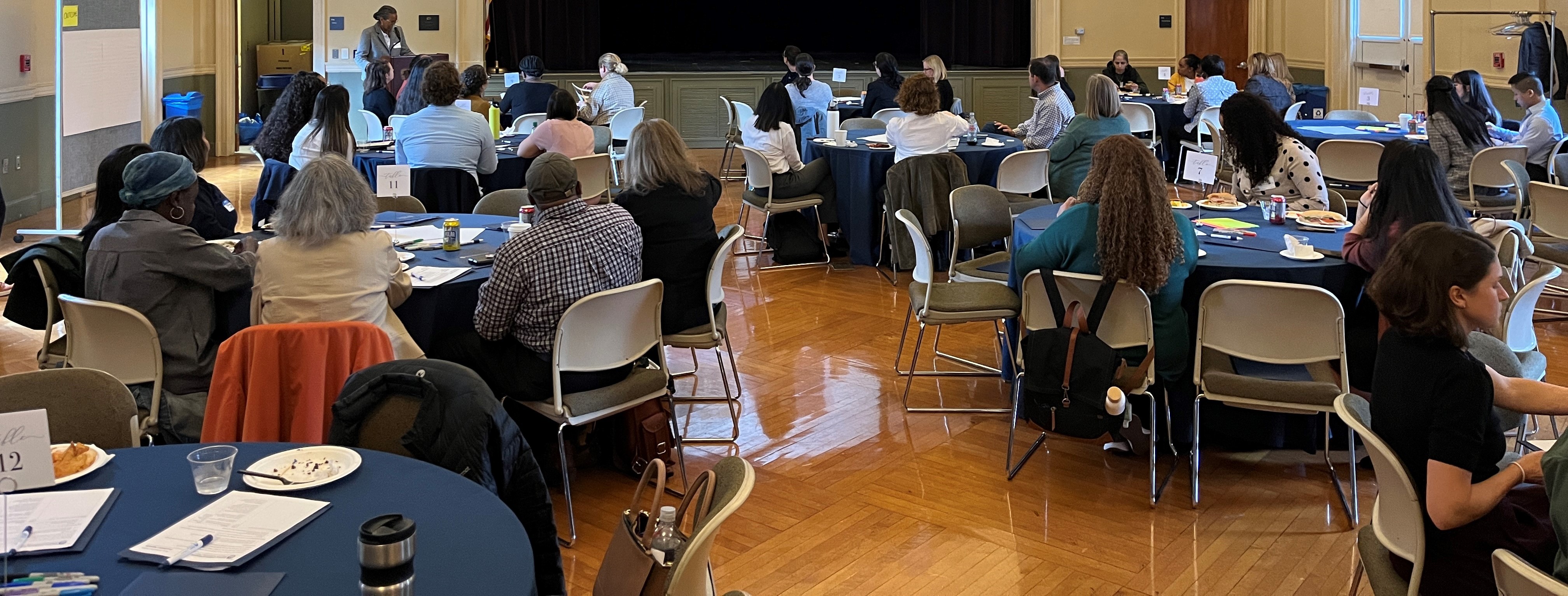 Woman speaking, people listening are seated at chairs around tables