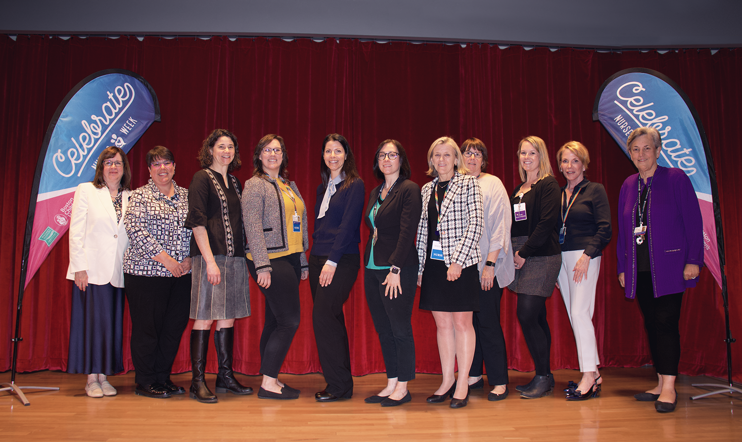 A group of nurses pose on stage in front of celebrate nurses week flags on either end of the stage.