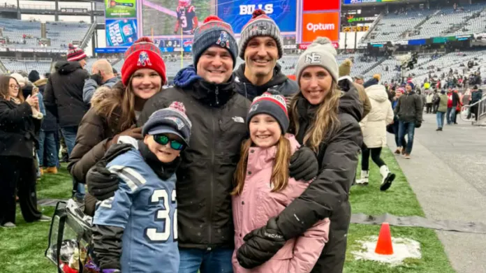 Three adults — two men and a woman — and three children stand on Gillette Stadium field with large scoreboard in background.