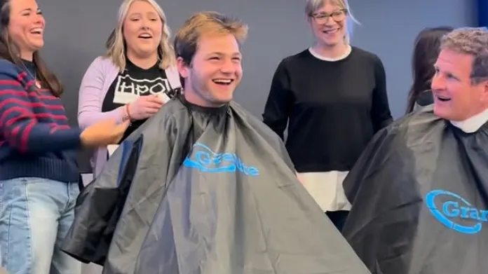 Young man sits in barber chair, covered in apron, laughing, while four other people in shot are also laughing