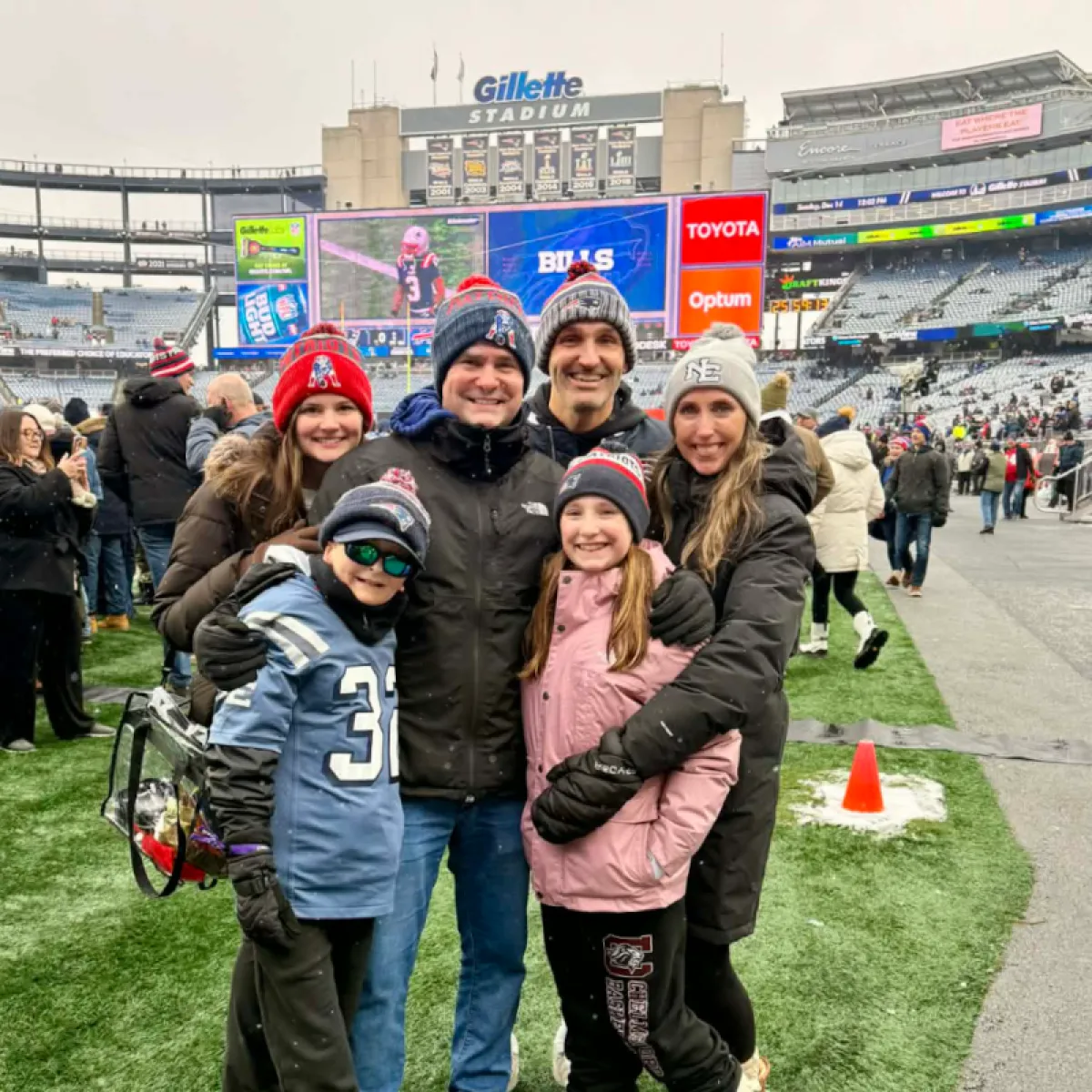 Three adults — two men and a woman — and three children stand on Gillette Stadium field with large scoreboard in background.