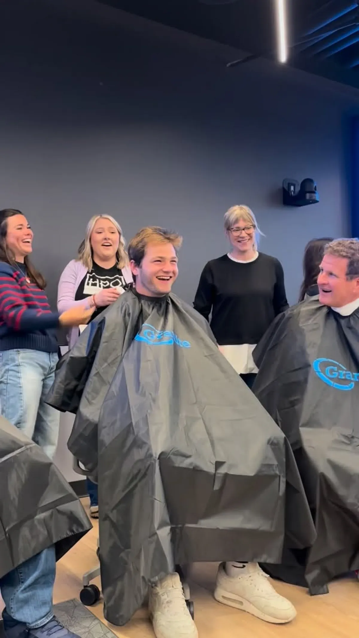 Young man sits in barber chair, covered in apron, laughing, while four other people in shot are also laughing