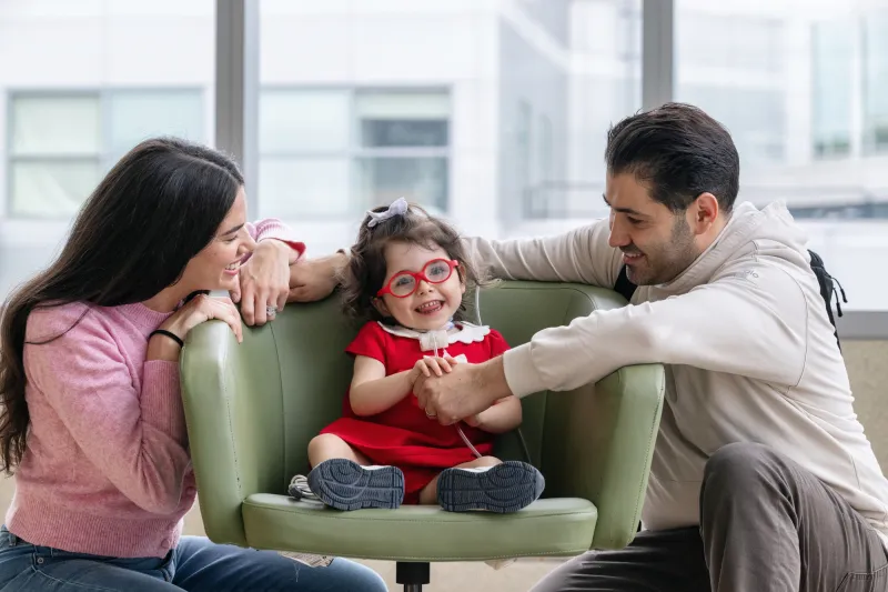 A smiling young child in red glasses sits in a green chair holding hands with two adults who are leaning in and smiling at her.