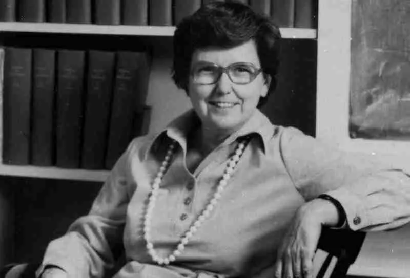 Woman wearing open-collar dress and necklace sits in front of a bookcase