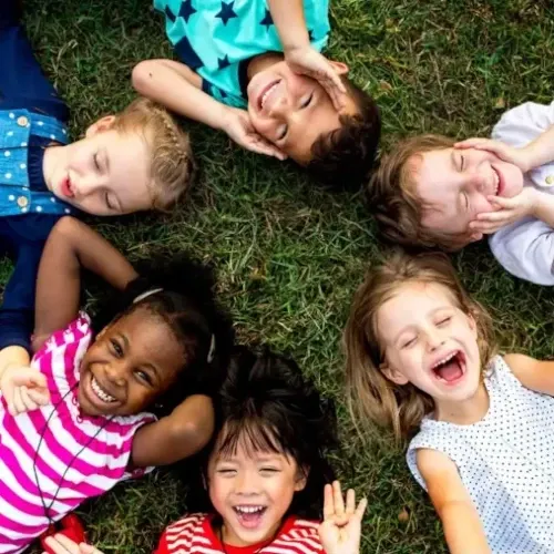 Six children lay on grass in a circle while making funny faces