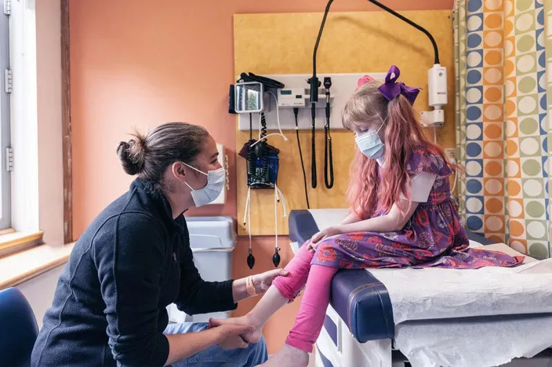 Female clinician examines lower leg of young girl patient. Both are wearing clinical-grade face masks.