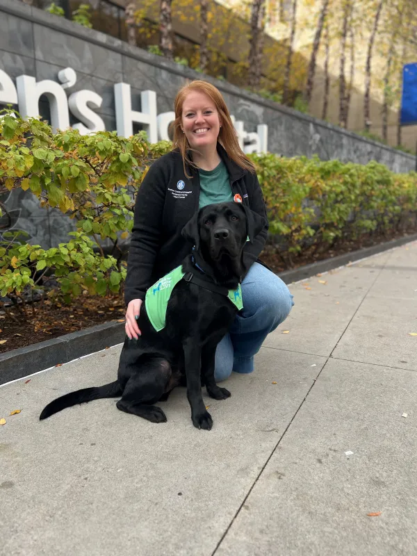 Dog rests on ground, with female handler on one knee, in front of Boston Children's Hospital sign