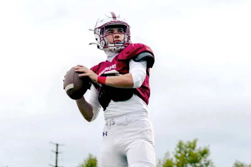 Quarterback, wearing helmet, prepares to throw pass with right hand, left hand holds onto football