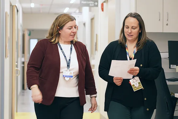 Two female clinicians walk down hospital corridor. Woman on left looks down at paper she holds. Woman on right looks on.