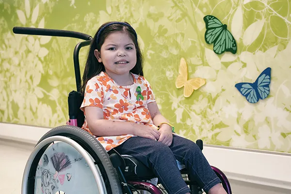 Girl sits in wheelchair in front of wall with painted mural featuring butterflies