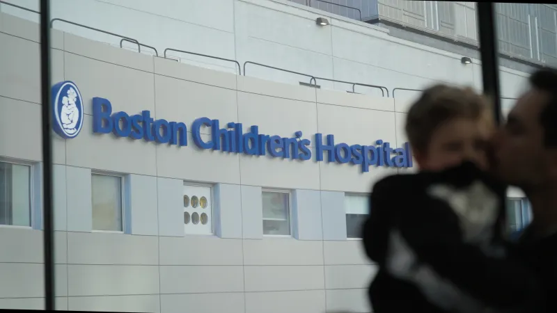 Adult holds small child at right of image; in background is large exterior signage for Boston Children's Hospital