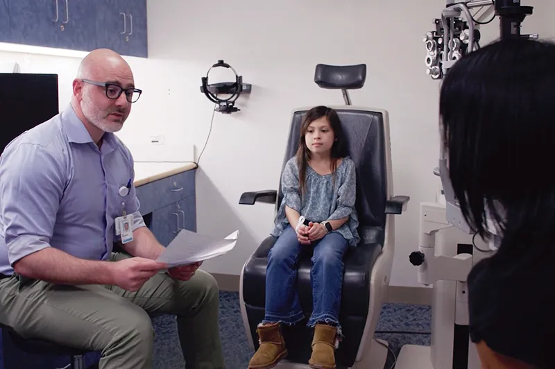 A doctor with paper in his hands while a child sits in the eye exam chair.