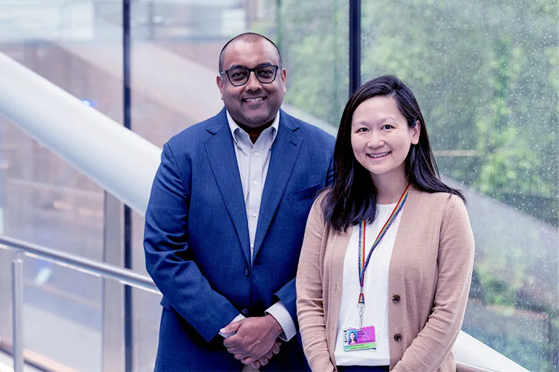 Man in blue blazer and blue shirt and woman in beige sweater and white shirt smile while standing on skybridge.
