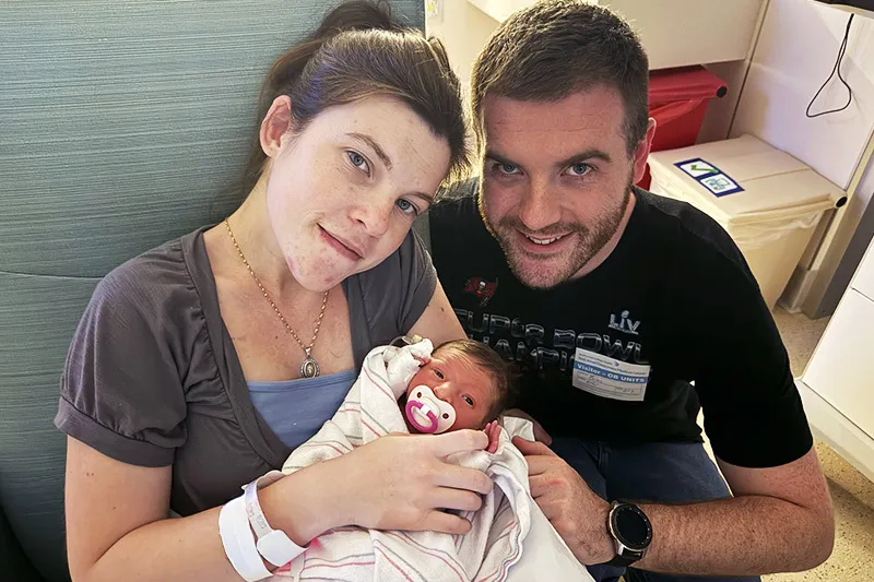 A mother who is seated in a hospital chair holds her infant daughter while the father leans in from the side.