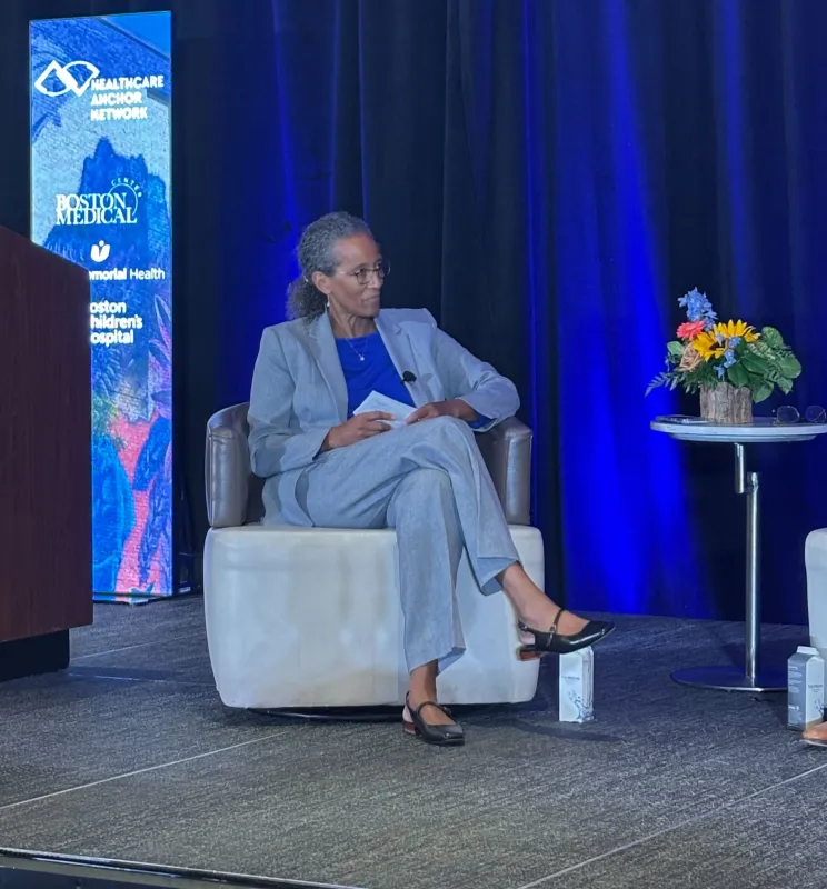 Woman with pulled-back hair and wearing glasses sitting on a stage speaks to unseen panelist during presentation