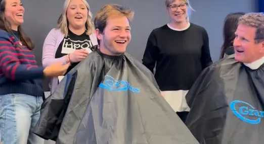 Young man sits in barber chair, covered in apron, laughing, while four other people in shot are also laughing