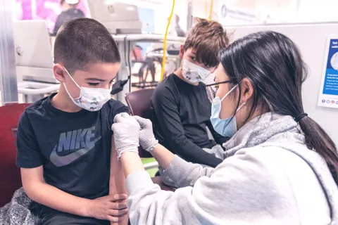 Woman clinician puts bandage on left arm of boy who is wearing a face mask, after an immunization