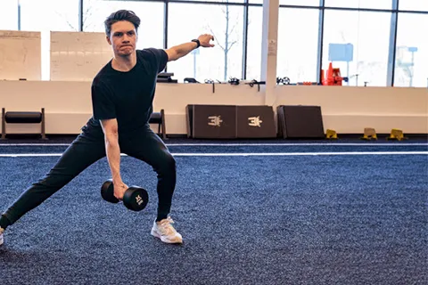 Young male holds dumbbell in right hand and does body stretches while standing on yoga mat